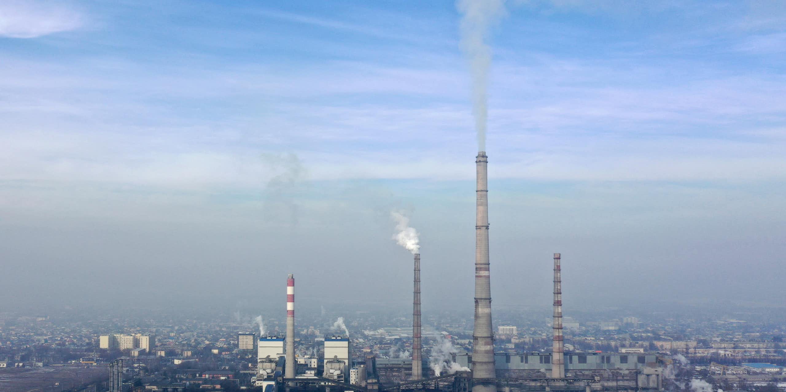 Tall facilities at a thermal power plant emit a gray substance over a city, with smog in the background.