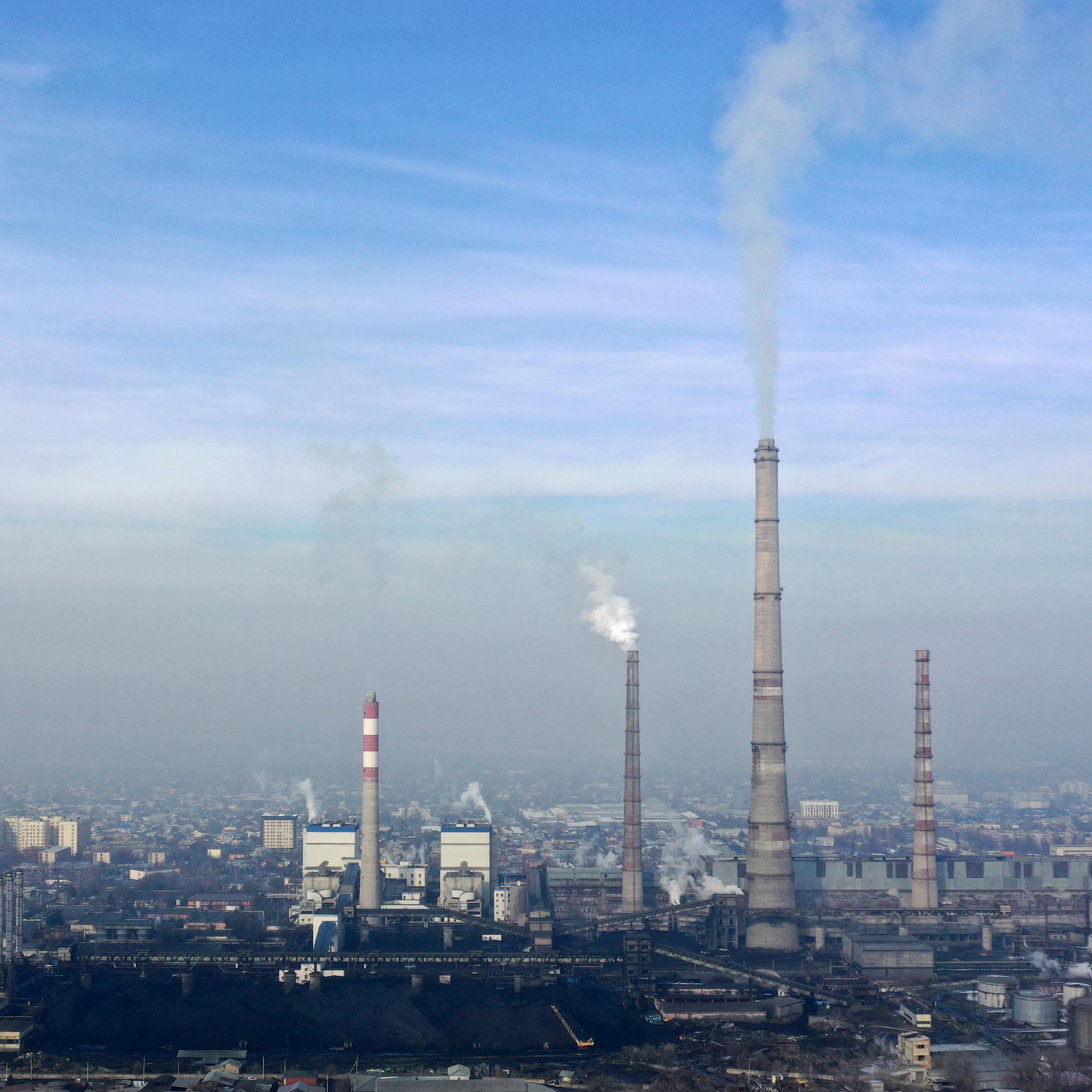 Tall facilities at a thermal power plant emit a gray substance over a city, with smog in the background.