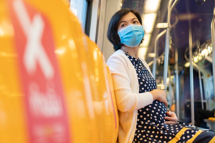A pregnant Asian woman wearing a mask sits on public transport.