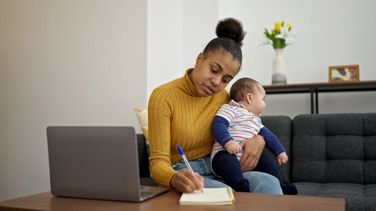 A black woman holds her infant baby while writing in a notebook and working on her laptop.