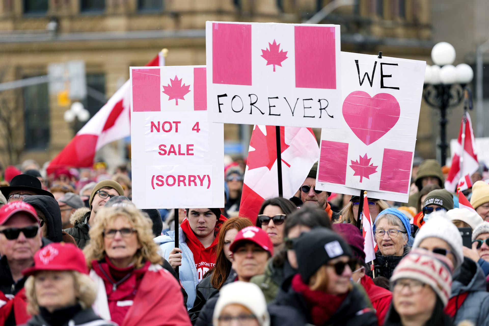 A crowd of protestors. Some of the people hold protest signs that state "Canada not 4 sale," "Canada Forever" and "We love Canada".