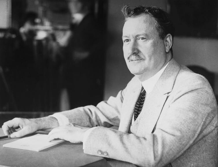 A white man with a mustache and a blazer sits at a desk with a paper in front of him in a black and white photo.