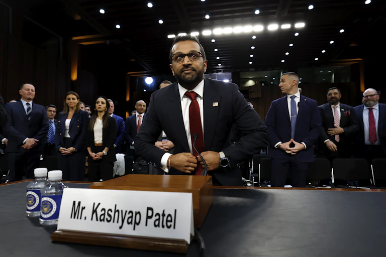 A brown skinned man wearing a black suit with a white shirt and red tie sits in front of a sign that says Mr. Kashyap Patel. A group of people formally dressed stand behind him.