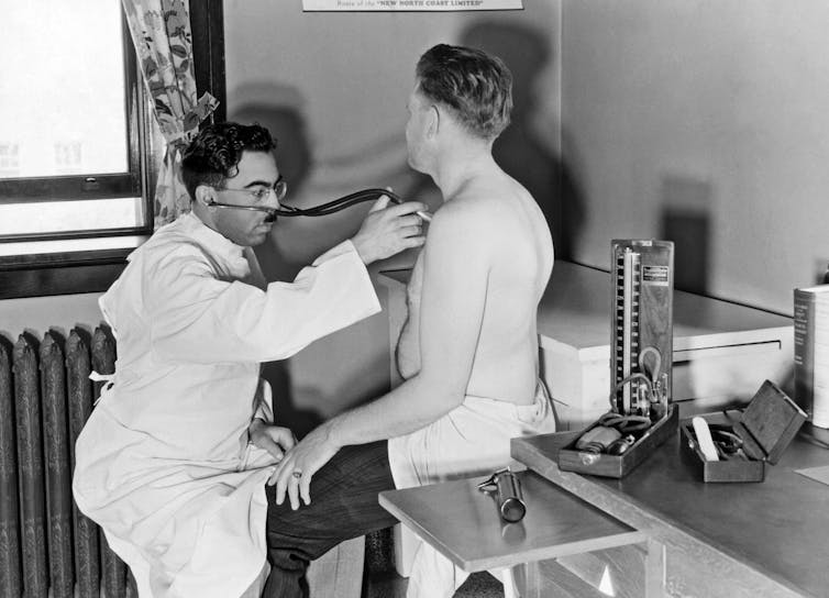 white-coated doctor uses stethoscope to listen to shirtless man's chest in exam room