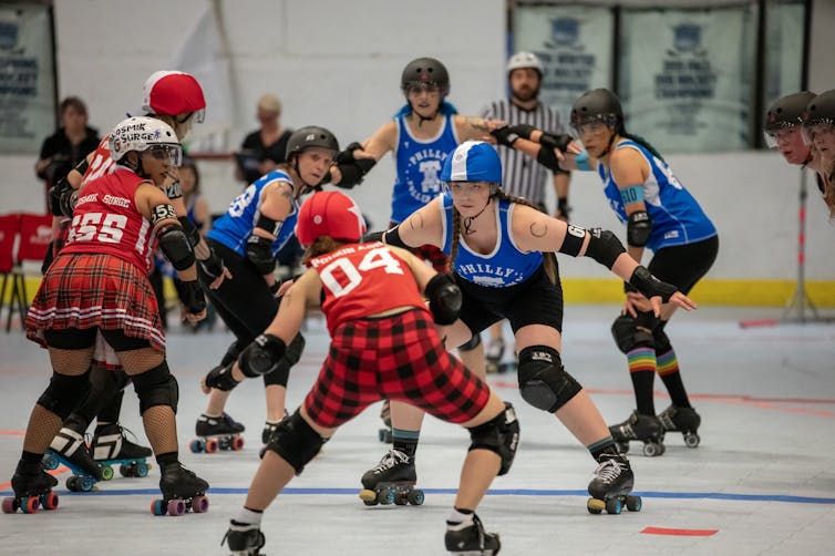 Three roller skaters in red uniforms face four skaters in blue uniforms with a referee in the background
