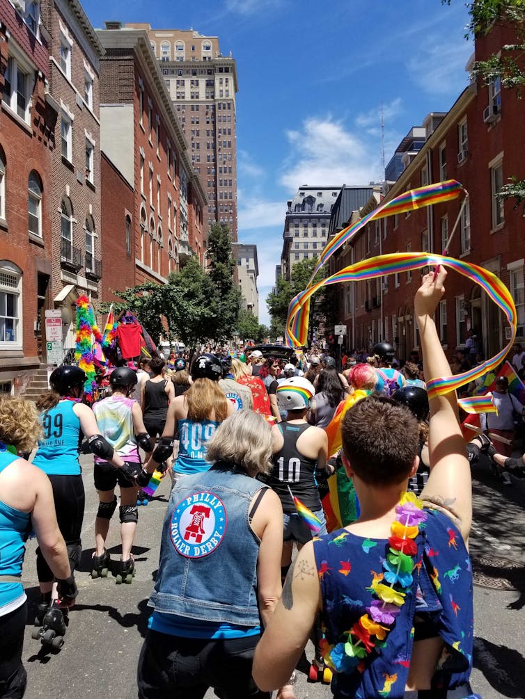 Skaters pictured from behind participate in a parade and wear rainbow accessories