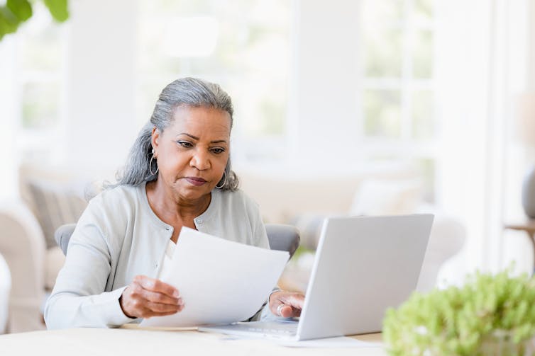 Woman with gray hair looking at paper with laptop open beside her