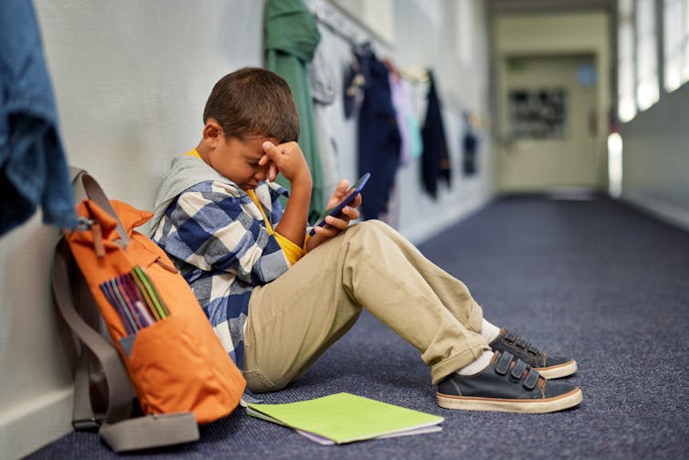A young boy sits on the floor of a school corridor with his hand on his head