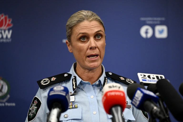 A woman in a police uniform stands behind a row of media microphones and speaks