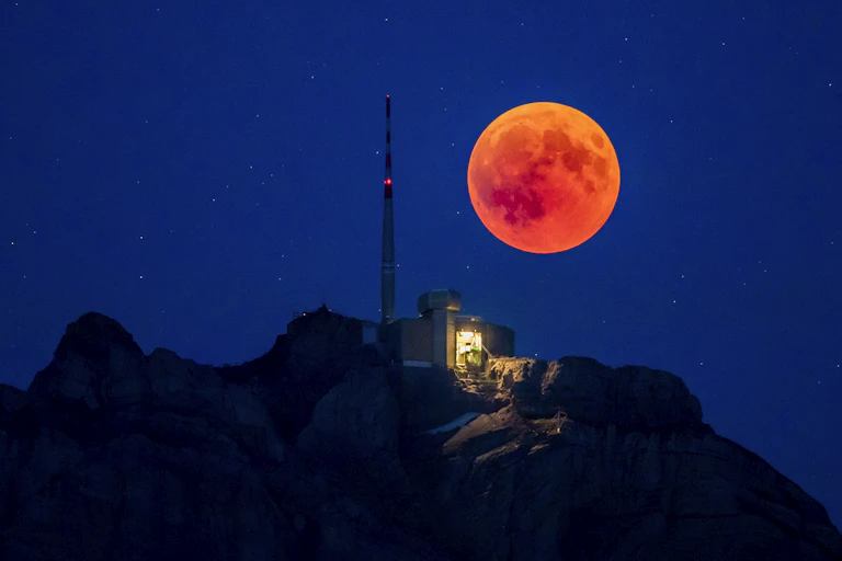 A full red moon above an illuminated radio hut on a hilltop.