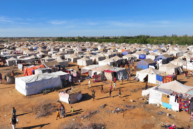 An aerial shot of a dusty field dotted with several white tents with several people walking around