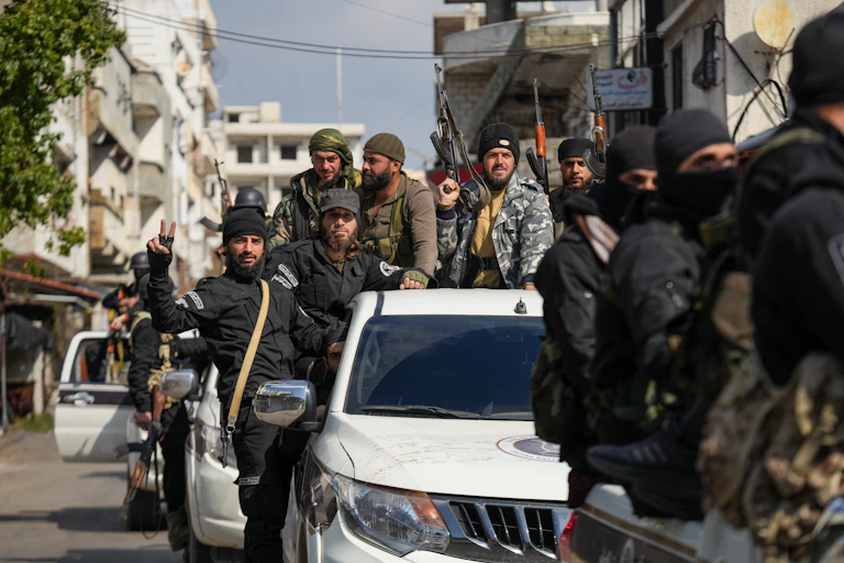 Armed Syrians in military uniforms on patrol in pick-up trucks. One is gesturing to the camera.