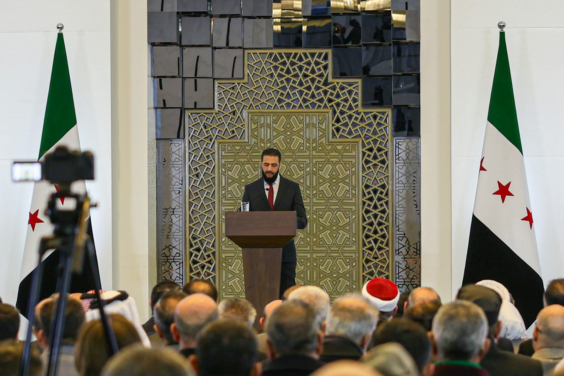 Syrian leader Ahmad al-Sharaa speaks to a large audience from a podium flanked by national flags, February 2025.