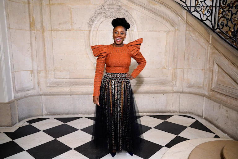 An African woman with an elaborate hairstyle smiles broadly as she poses near a vintage staircase, wearing a black skirt and an orange blouse with winged shoulders.