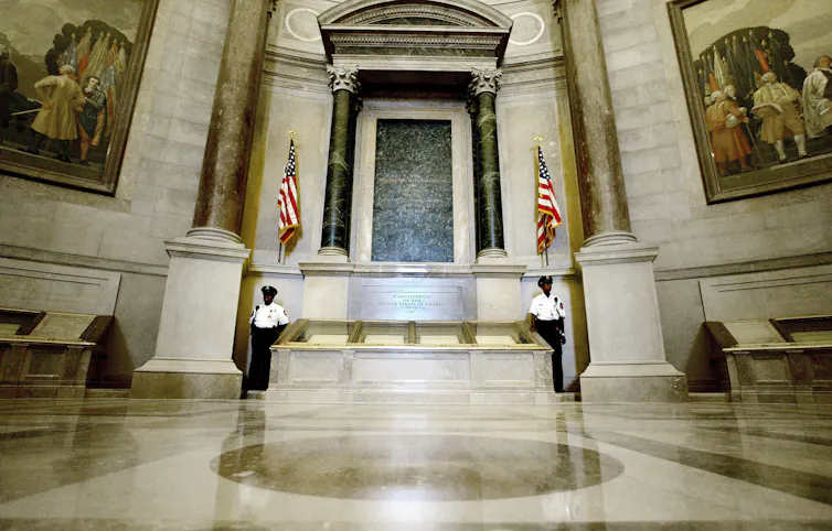 Two guards stand next to a case that contains the U.S. Constitution in an ornate rotunda.