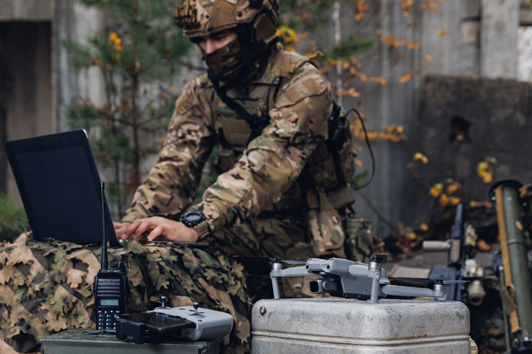 A soldier in uniform types on a laptop with military hardware in the foreground.