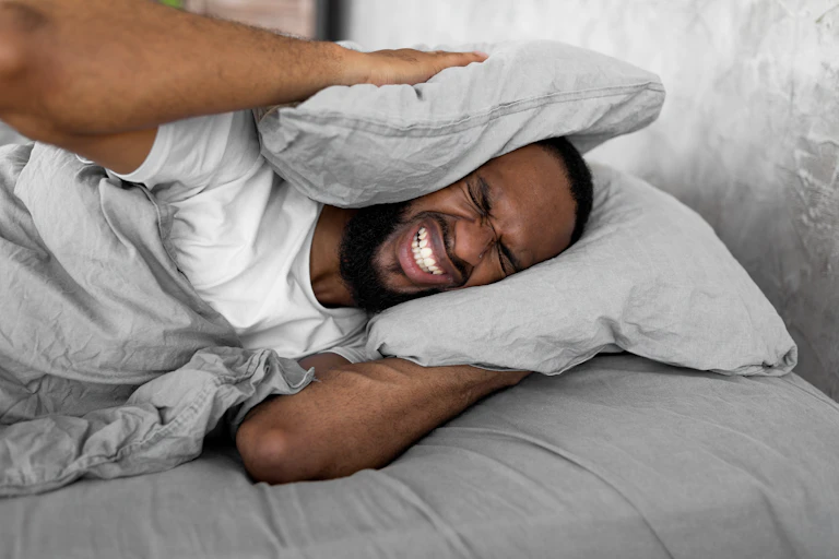 Um jovem tem dificuldade para dormir devido ao barulho. Ele está segurando dois travesseiros sobre os ouvidos, irritado.