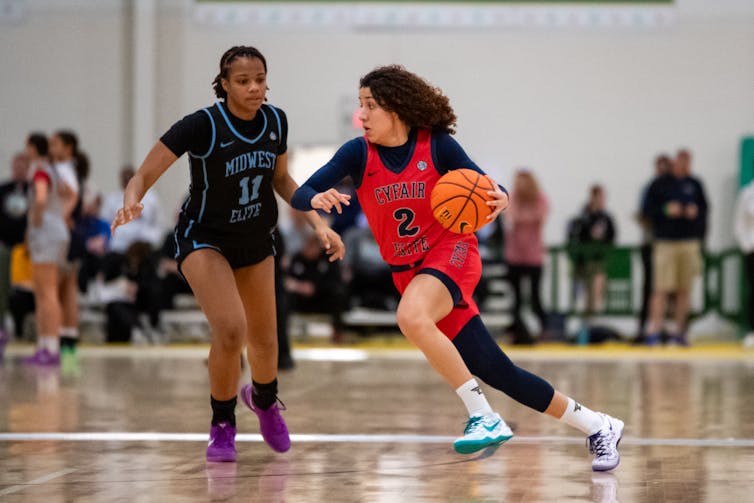 young woman wearing a red jersey dribbles a basketball on a court as she moves around another young woman wearing black
