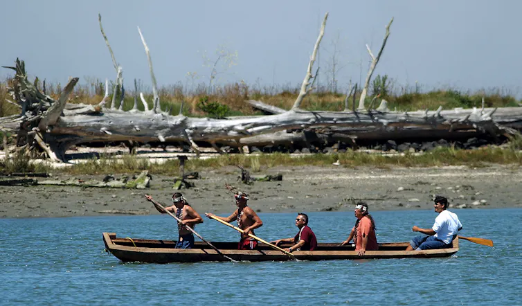 Five Native men paddle in a dugout, with an island in the background