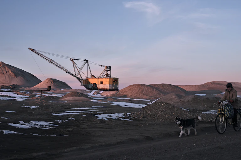 A person on a bicycle with a dog rides past an open mining area with a large excavator in the distance.