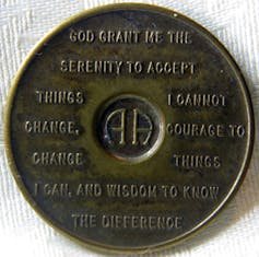 A small bronze disk with 'AA' in the middle, inscribed with a prayer in all capital letters.