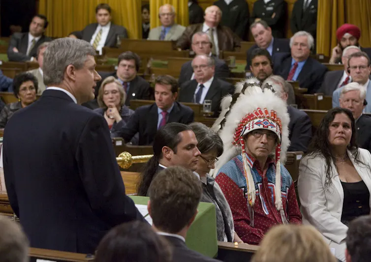 A man in a suit speaking as people listen, one in a white suit and one wearing regalia.