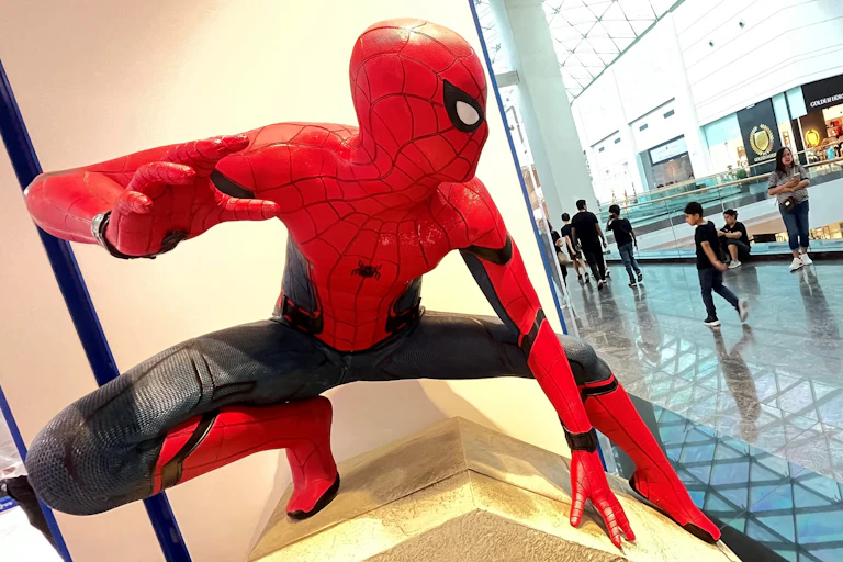 A statue of Spider-Man, positioned by a large window in a high-rise, shows him poised to jump as people walk below.