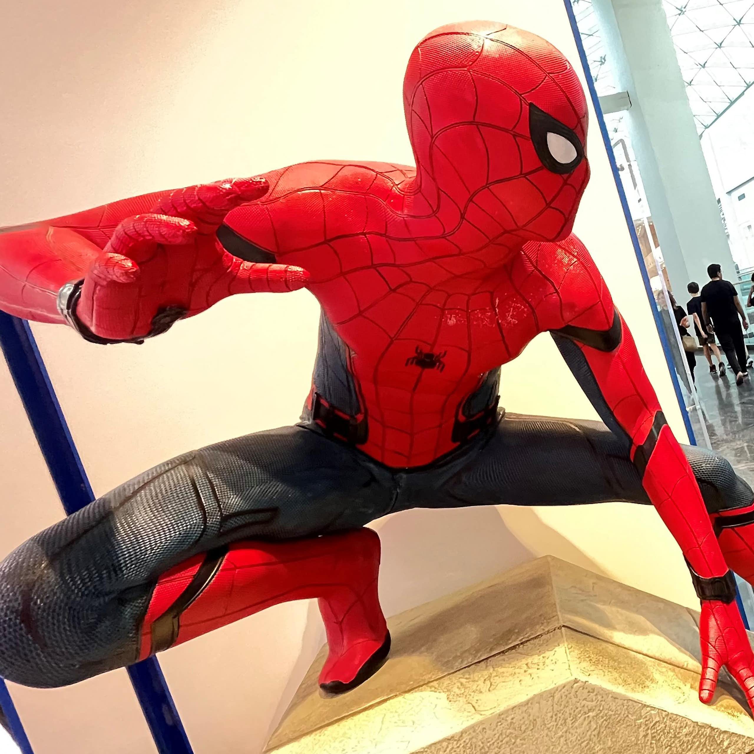A statue of Spider-Man, positioned by a large window in a high-rise, shows him poised to jump as people walk below.