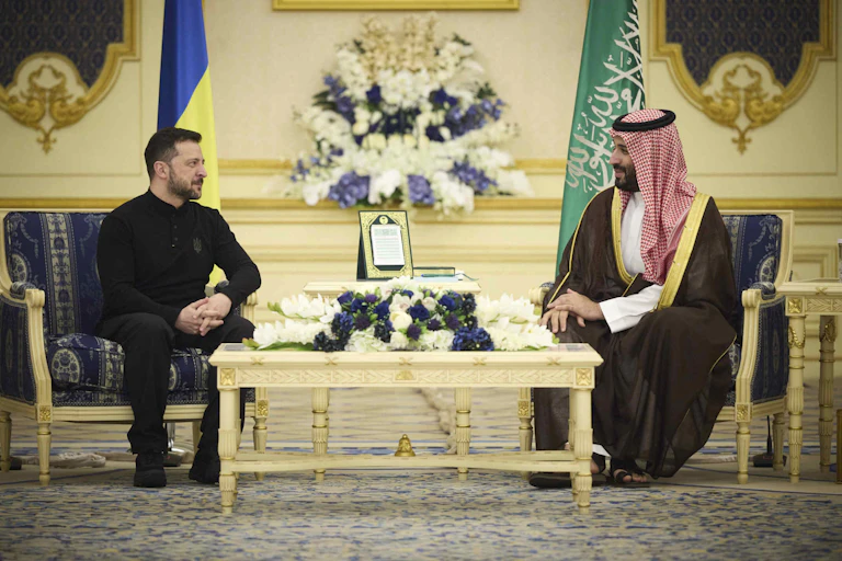 A dark-haired man with a beard in a dark shirt and pants talks to another man wearing robes and a red-and-white keffiyeh.