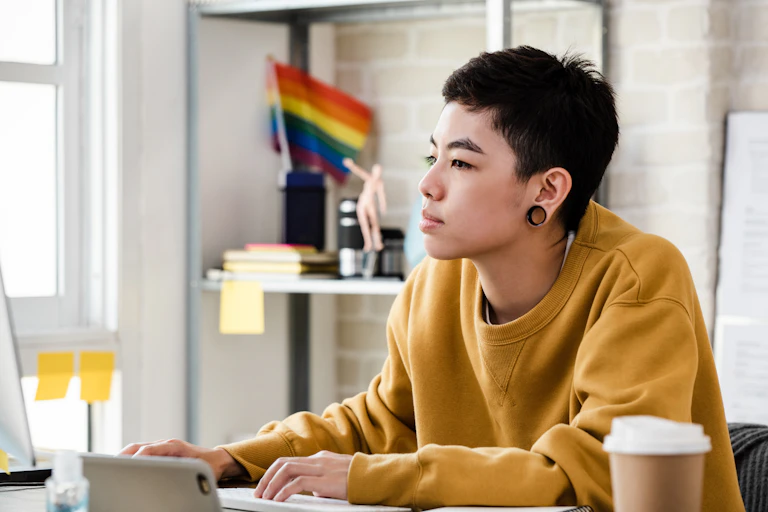 Young Asian tomboy woman in casual attire working from home in living room with Pride flag in background