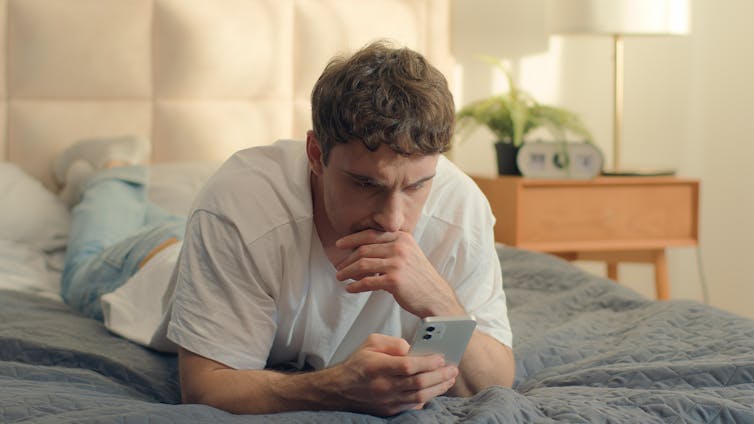 Worried man lies on his bed looking at phone.