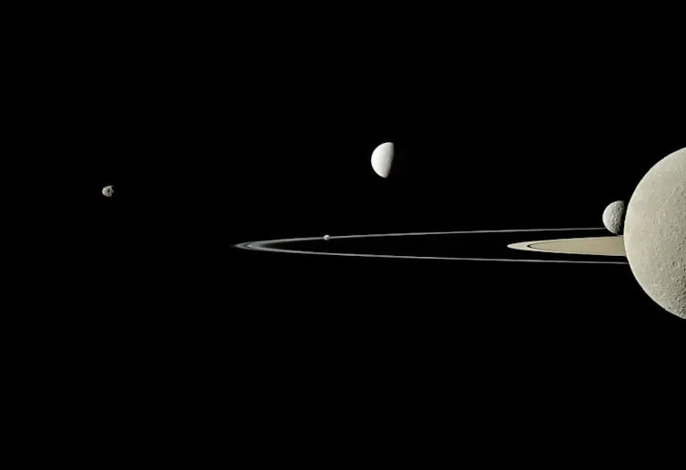 Uma imagem em escala de cinza mostrando Saturno e cinco de suas luas. Saturno está na extrema direita e metade dele não aparece na imagem. Os anéis são estreitos e se estendem diretamente do centro de Saturno na imagem até um pouco mais da metade da imagem em direção a