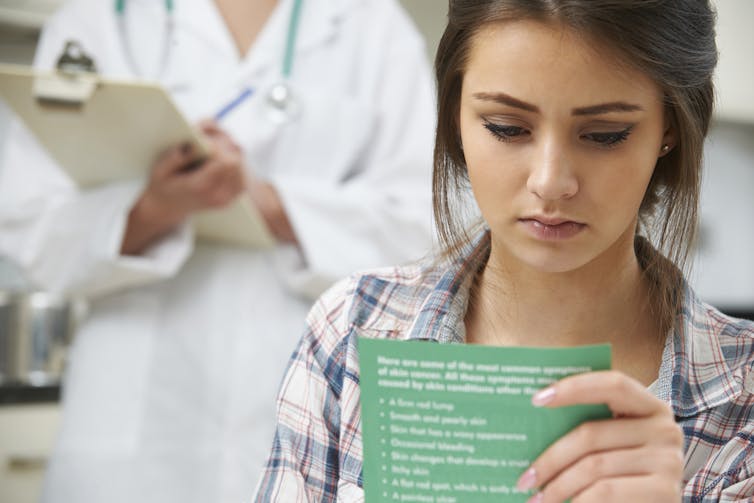 Young female patient reading information leaflet, doctor wearing white coat standing behind her