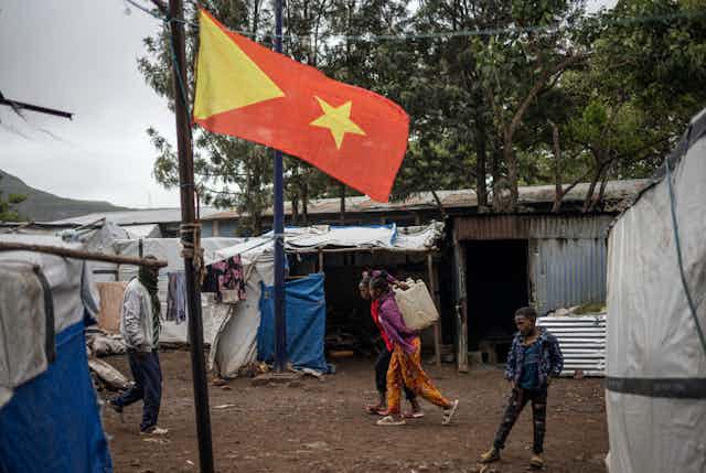 Three children and a man walking past makeshift shelters made with poles and white and blue canvas near a pole with a yellow and red flag