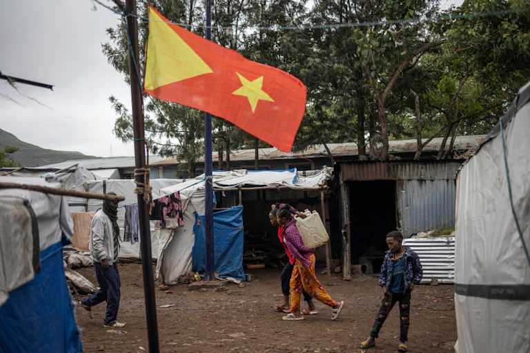 Three children and a man walking past makeshift shelters made with poles and white and blue canvas near a pole with a yellow and red flag