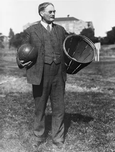 A black and white image of man in a 1930s suit holding a basketball and a basket.