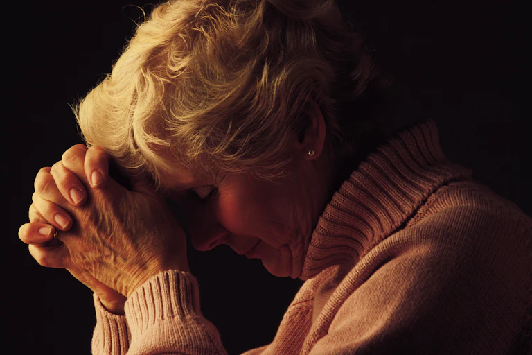 A woman with short white hair, wearing a pale pink turtleneck sweater, rests her head on her hands as if in prayer.