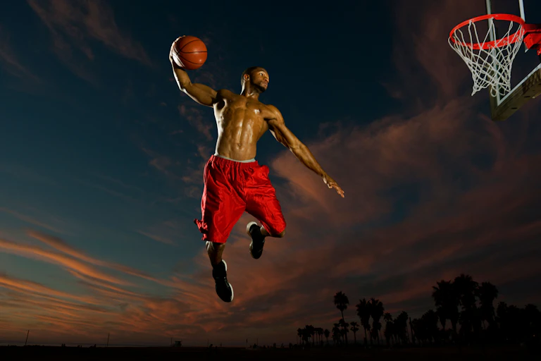 A shirtless black man in red shorts shooting at a basketball hoop.