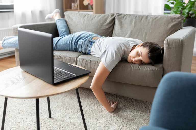 A young woman lying face down on a sofa with her laptop open on the coffee table in front of her