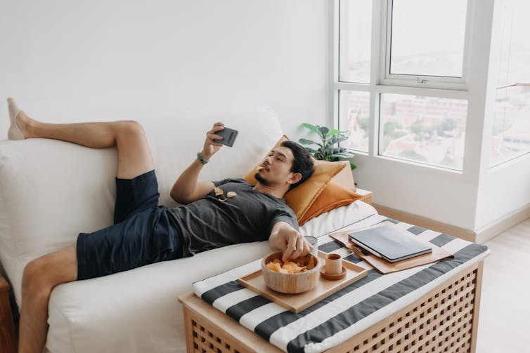 A man lying on a couch, messily eating potato chips and watching a video on his mobile phone