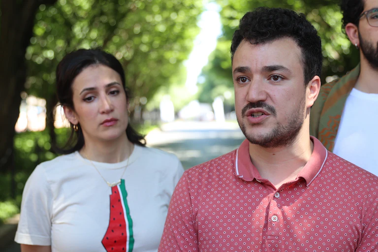 A man with black hair and a mustache stands in between two other young people with dark hair, with green trees behind them.