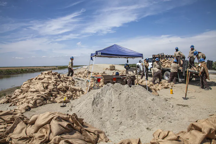 People load bags full of sand and stack them next to a body of water.