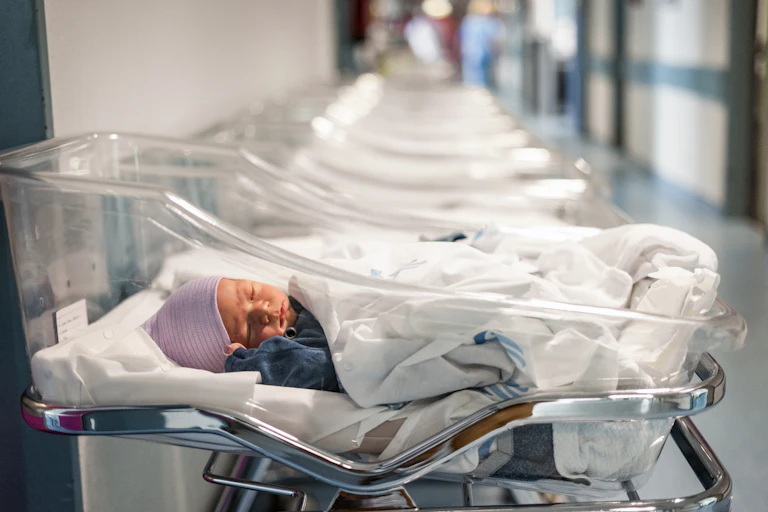 Dans un hôpital, un nouveau-né coiffé d'un bonnet rose est allongé dans un couffin en plastique transparent. Une rangée de berceaux similaires et vides sont disposés à côté de lui.