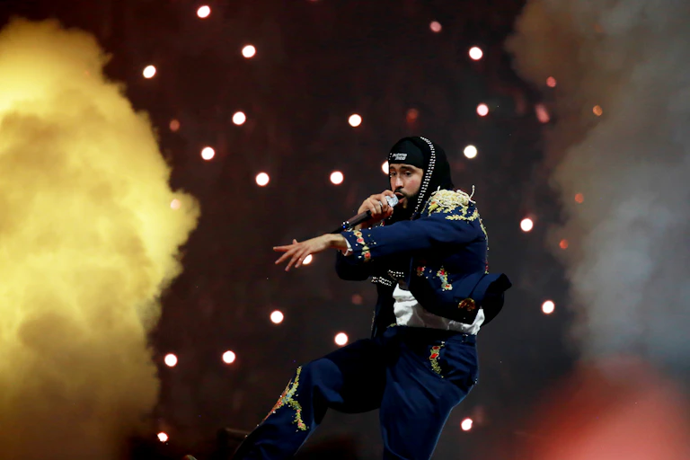 A young man wearing dark blue pants and jacket covered with appliqué flowers gestures on stage, holding a microphone; behind him is smoke and a spangle of stage lights.