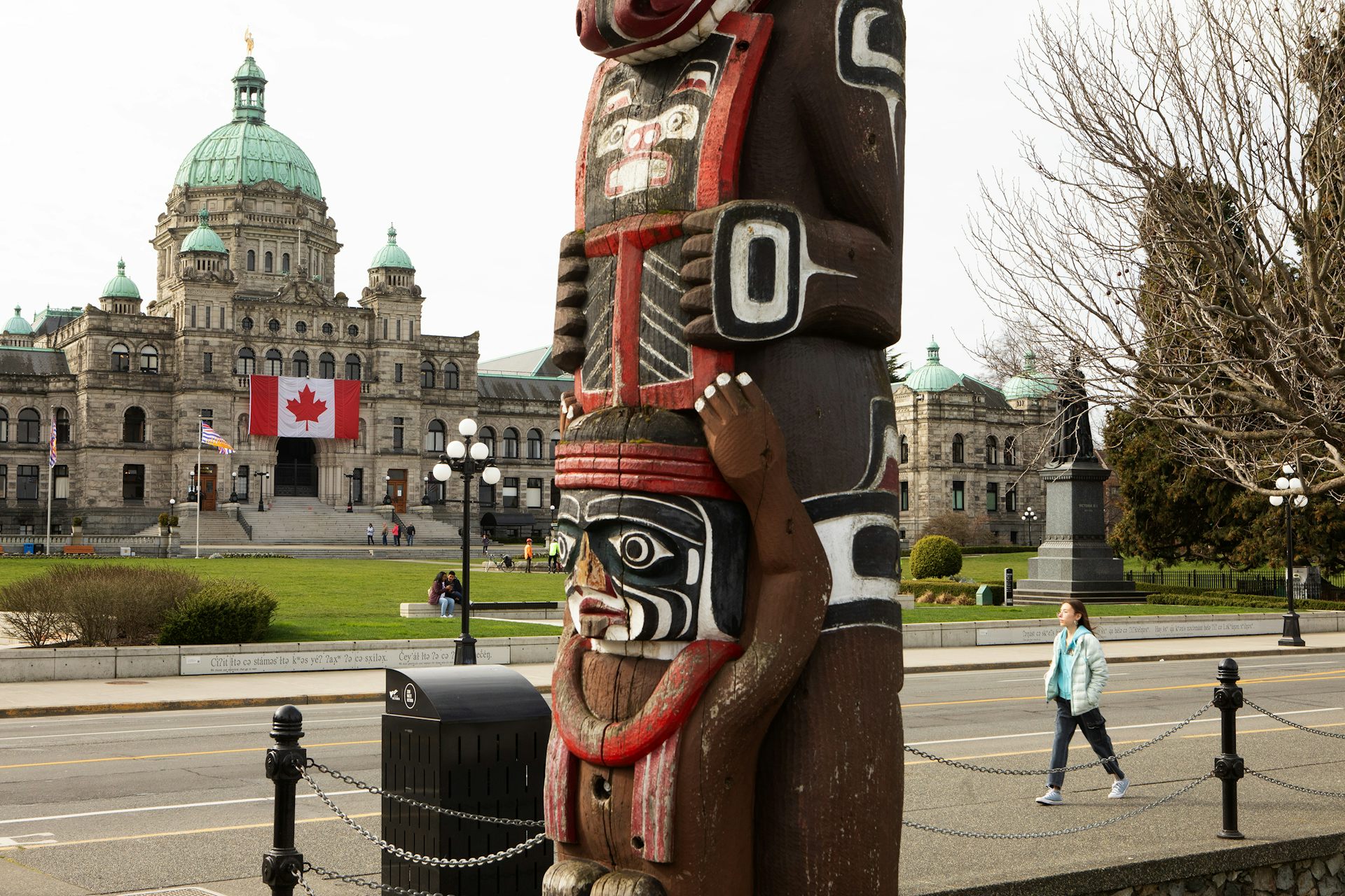 A totem pole is seen in the foreground, while a domed building with a Canadian flag over it is seen in the background 