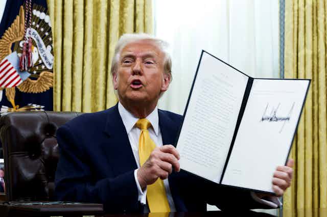 An old man with orange-tinged skin and whiteish hair wearing a dark suit and yellow tie holding a document with an oversized signature