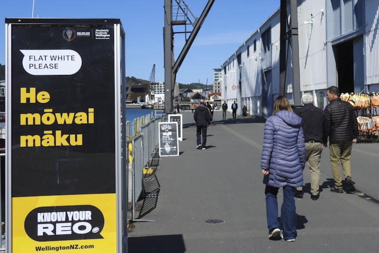 Des gens passent devant un panneau indiquant « flat white please » He mowai maku