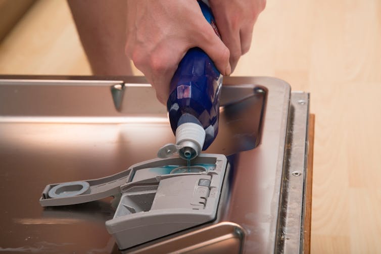 A person pours blue rinse aid into the dishwasher compartment.