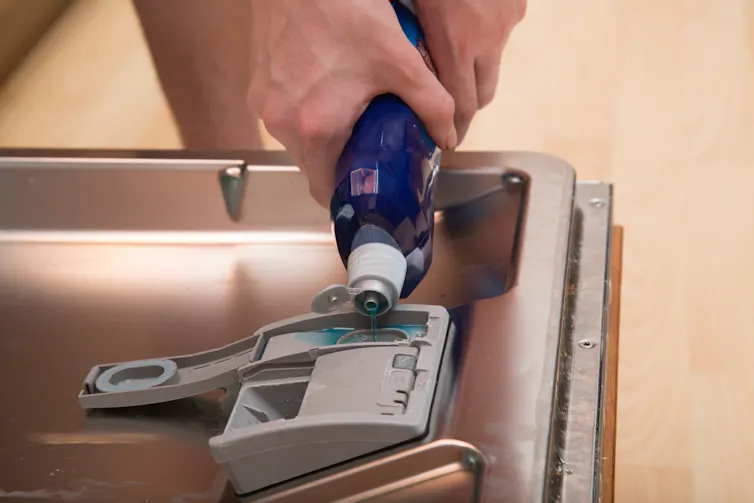 A person pours blue rinse aid into the dishwasher compartment.