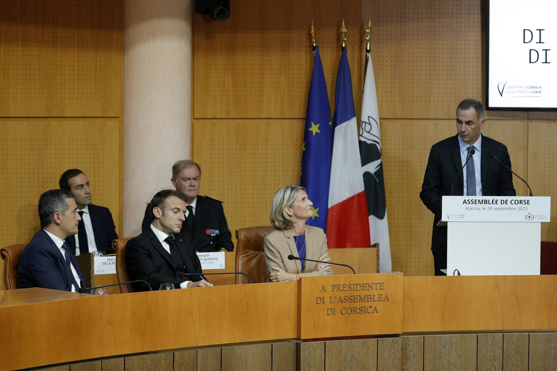 On voit un homme debout, le président du conseil exécutif de Corse Gilles Simeoni prononçant un discours devant le président Macron et le ministre français de l'Intérieur Gérald Darmanin qui l'écoutent.